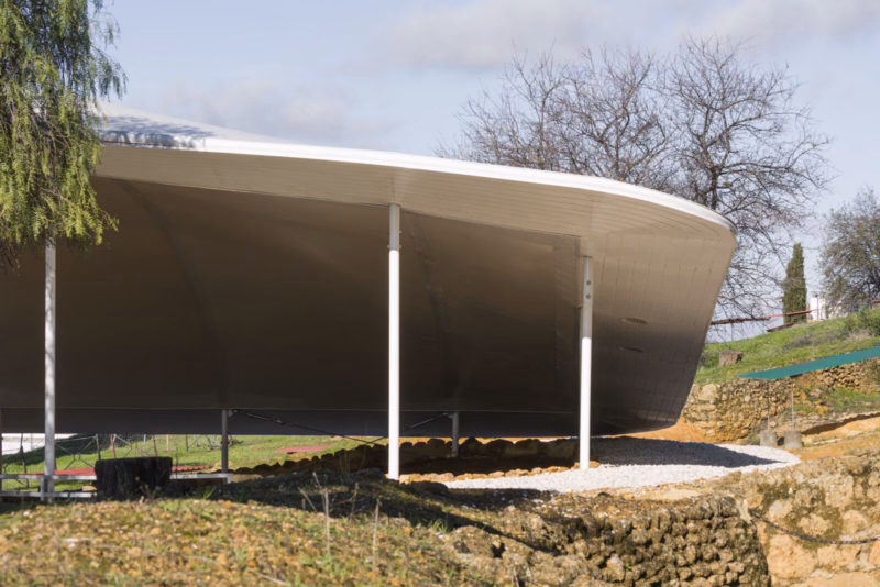 Low-angle side view of the textile canopy showing the curved double membrane and slender supports on the natural terrain. Nd_Arquitectos. Carmona, Spain. Photo by Jesús Granada.